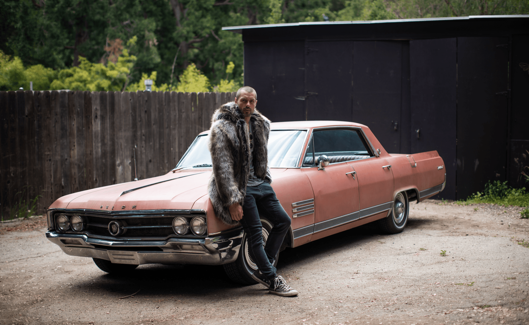 A man in a faux fur coat is leaning against a vintage car.