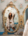 The reflection of a brunette female model with one hand on top of her head, slightly tilted back, appears in a gold-framed mirror against an ornate, wallpapered wall. She is wearing a realistic faux fur Siberian snow leopard coat.