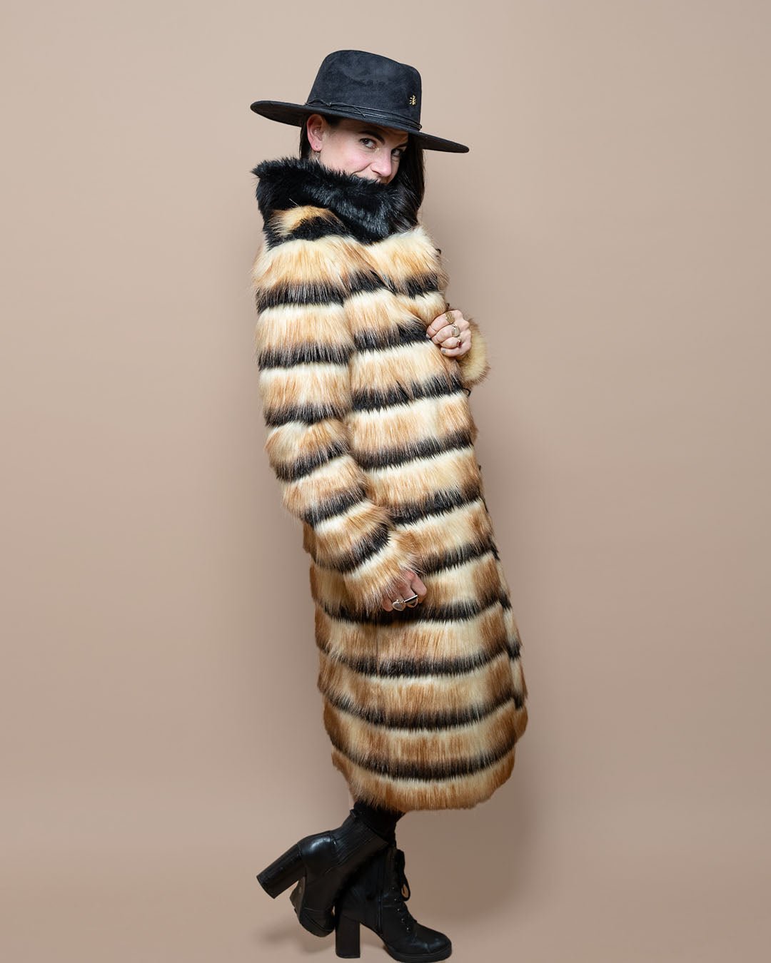 Model in black fedora, collar up covering mouth, looking over Long Gazelle Vegan Fur Coat held closed to showcase full style.