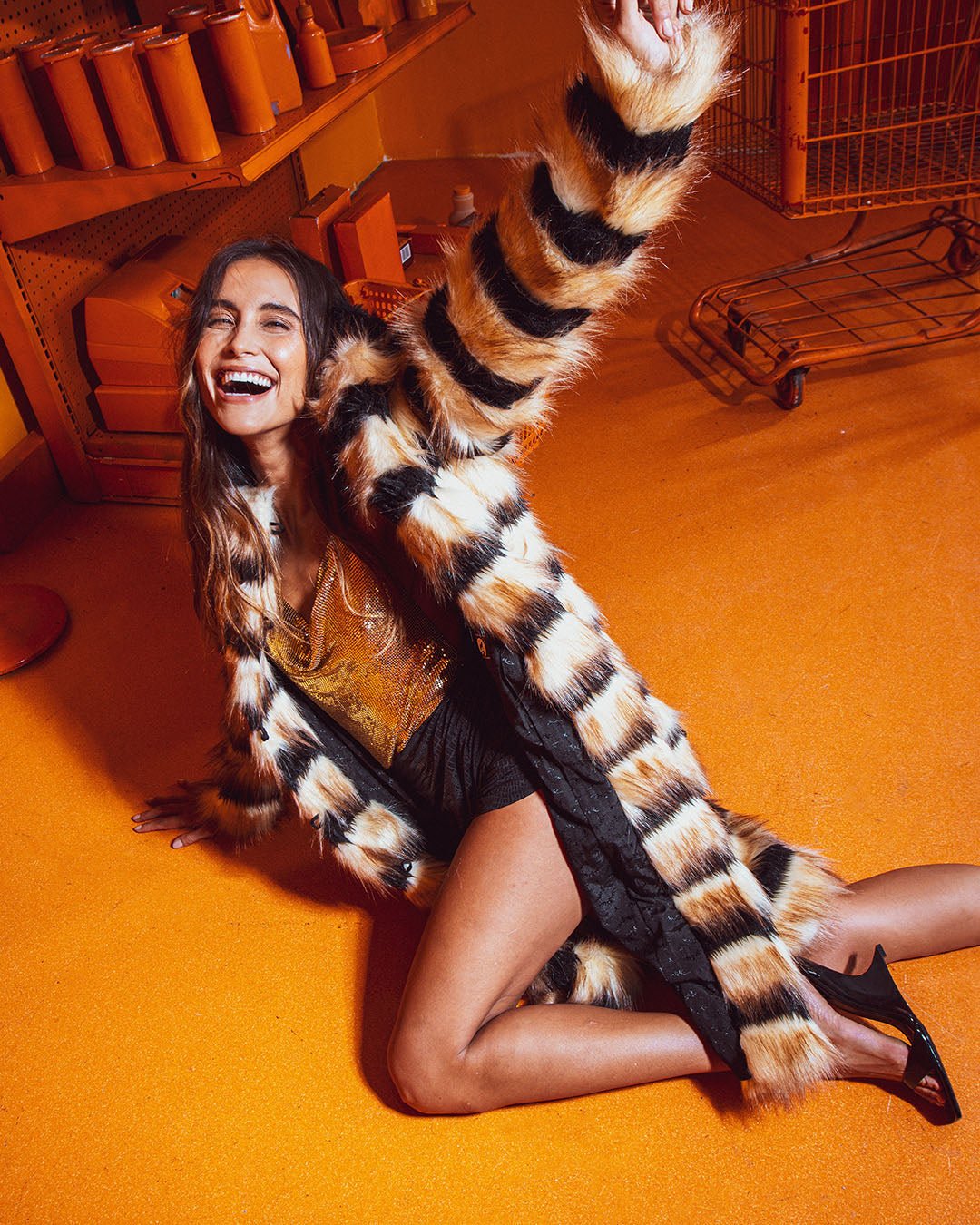 Laughing model in orange room, playfully on floor with one hand outstretched, legs bent, in Long Collared Gazelle Faux Fur Coat.