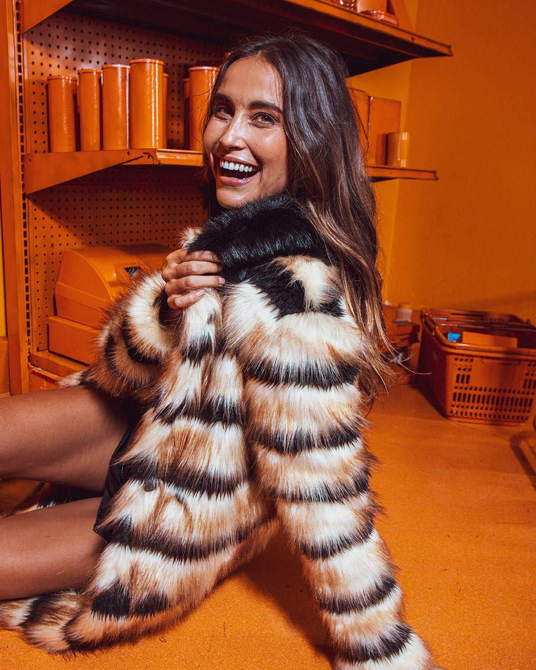 Laughing brown-haired model leaning back on one hand in orange room, other hand holding the Collared Gazelle Long Faux Fur Jacket.