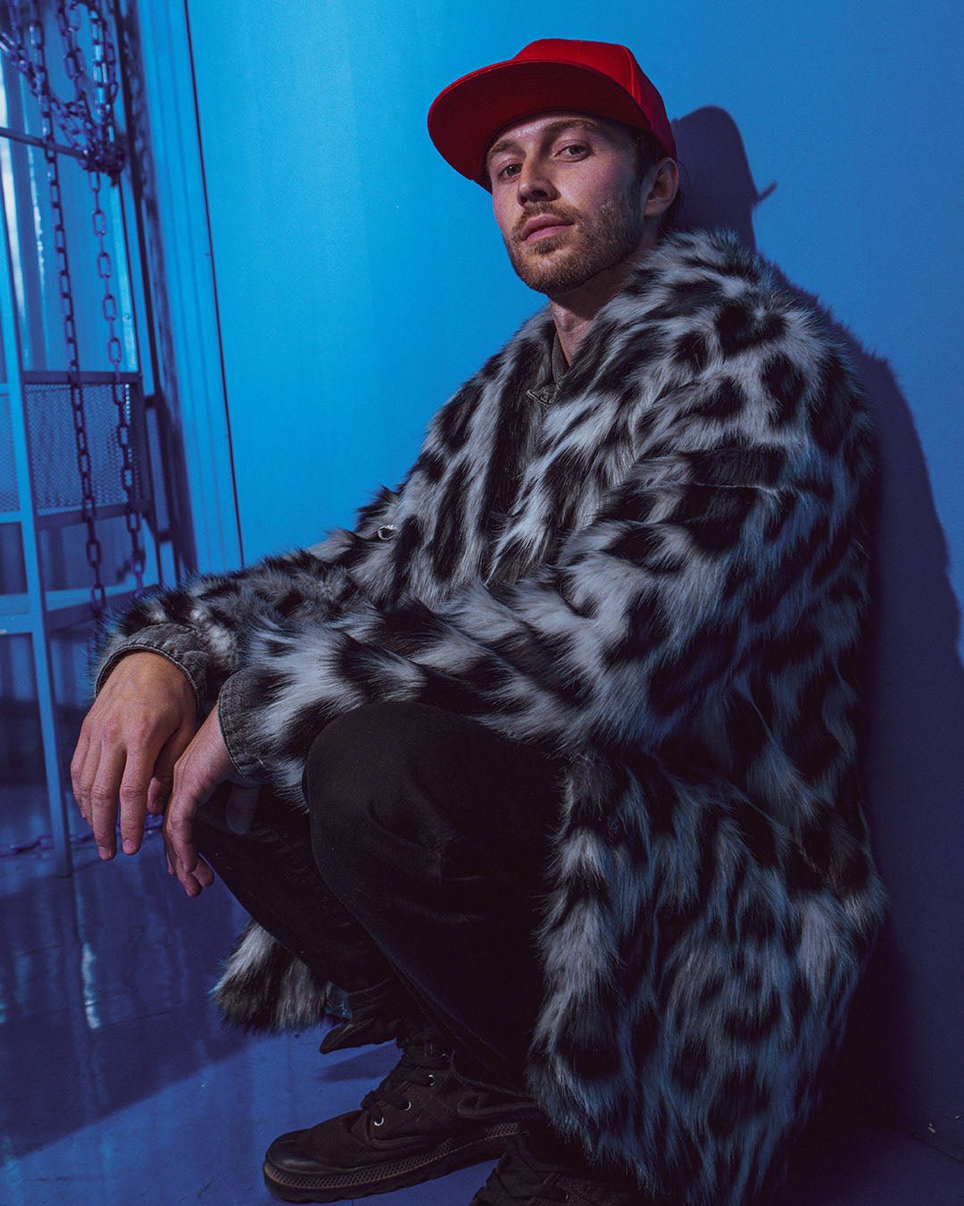 Man squats in an all-blue room, hands over knees, wearing the Collared Himalayan Snow Leopard Faux Fur Coat. Edgy and cool.