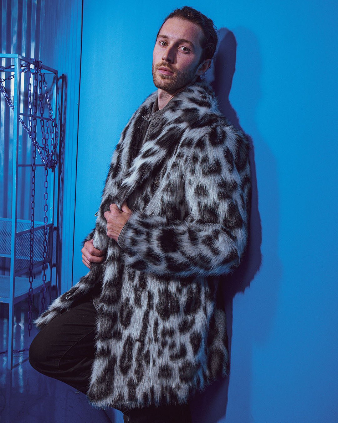 Guy leans against blue wall, knee bent, holding lapels of Himalayan Snow Leopard Faux Fur Jacket. Stylish and edgy.