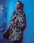 Guy leans against blue wall, knee bent, holding lapels of Himalayan Snow Leopard Faux Fur Jacket. Stylish and edgy.