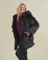 Blond gal with curves side facing and  smiling with a hand in the pocket of vegan black wolf fur coat with hood down.