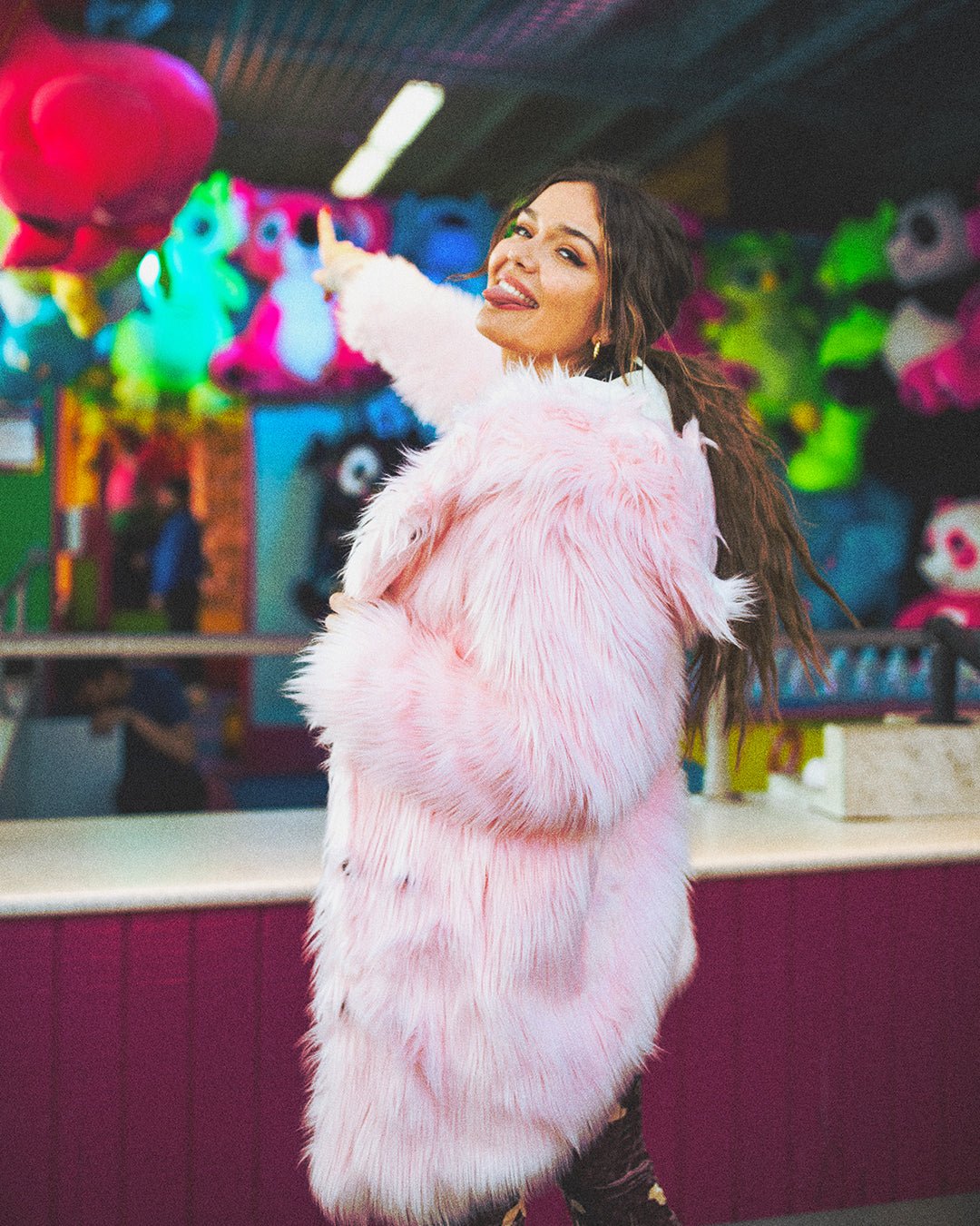 Girl at a carnival booth sticking her tongue out and pointing at her prize, wearing the Flamingo Wolf faux fur jacket with the hood down.