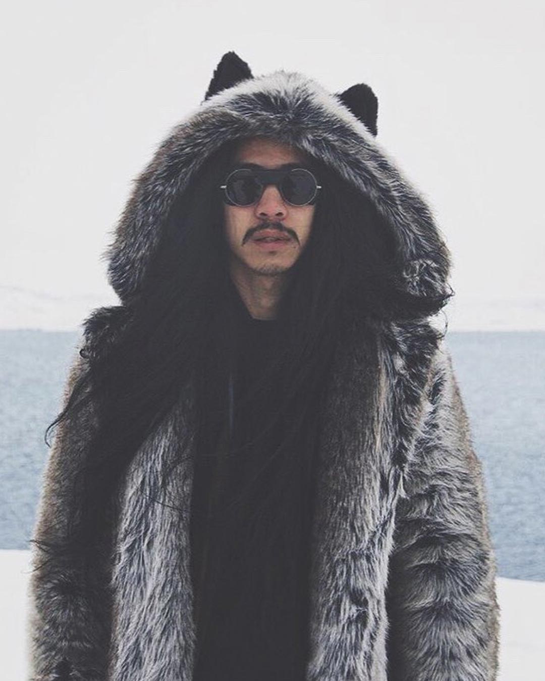 Man with long dark hair wearing sunglasses and a hooded grey faux fur coat.