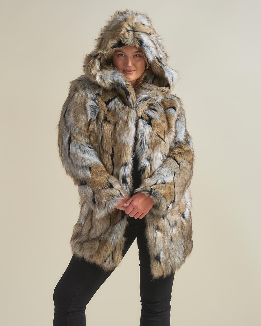 A demure model wearing a Hooded Wolverine fake fur jacket with the hood up, clasping her hands in front of her and looking directly at the camera.