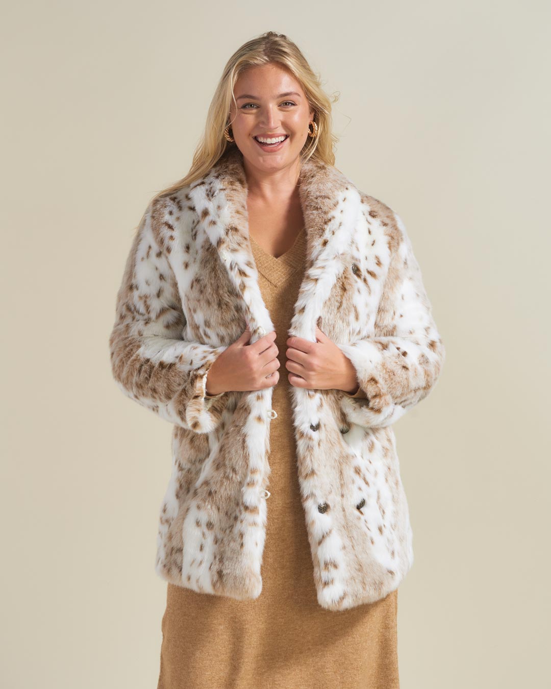 A blonde, curvy beauty is smiling and holding the bottom lapels of a Siberian snow leopard faux fur coat with a collar that she’s wearing.