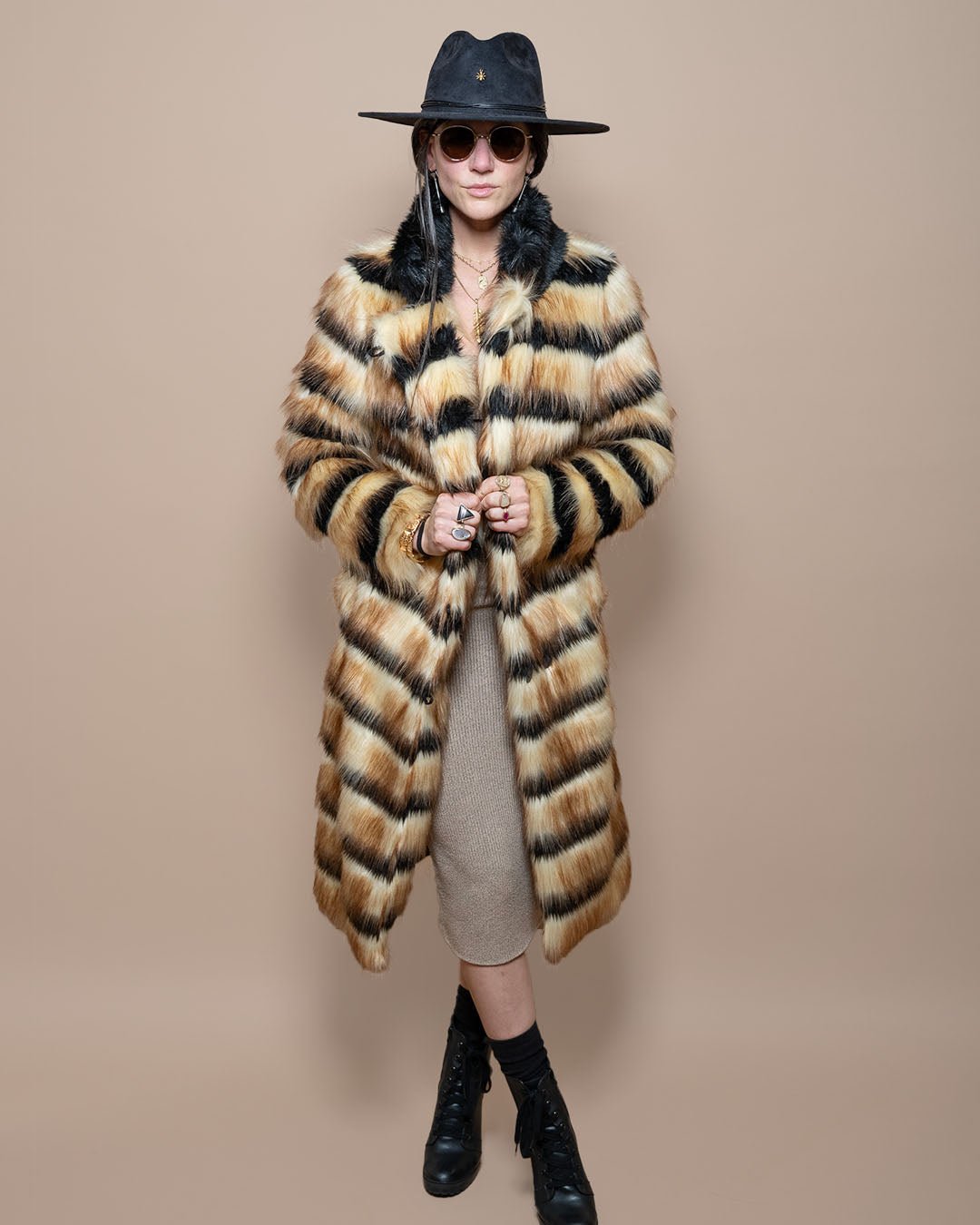 Dark-haired model in black fedora and sunglasses, holding lapels of Long Collared Gazelle faux fur coat, ankle-crossed pose.