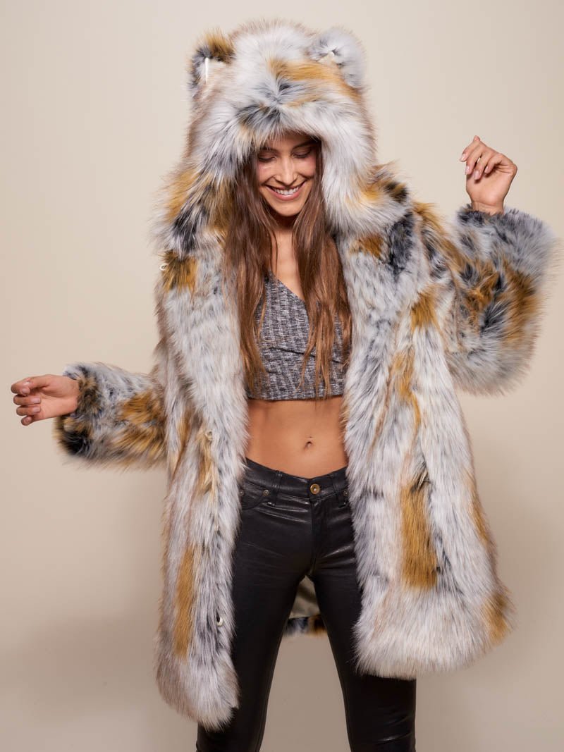 A gal in an all-orange room, leaning on an orange shopping cart with one hand, the other hand on the top of her hood with ears, wearing orange sunglasses and the Arctic Fox Classic faux fur coat, with one leg crossed over the other.