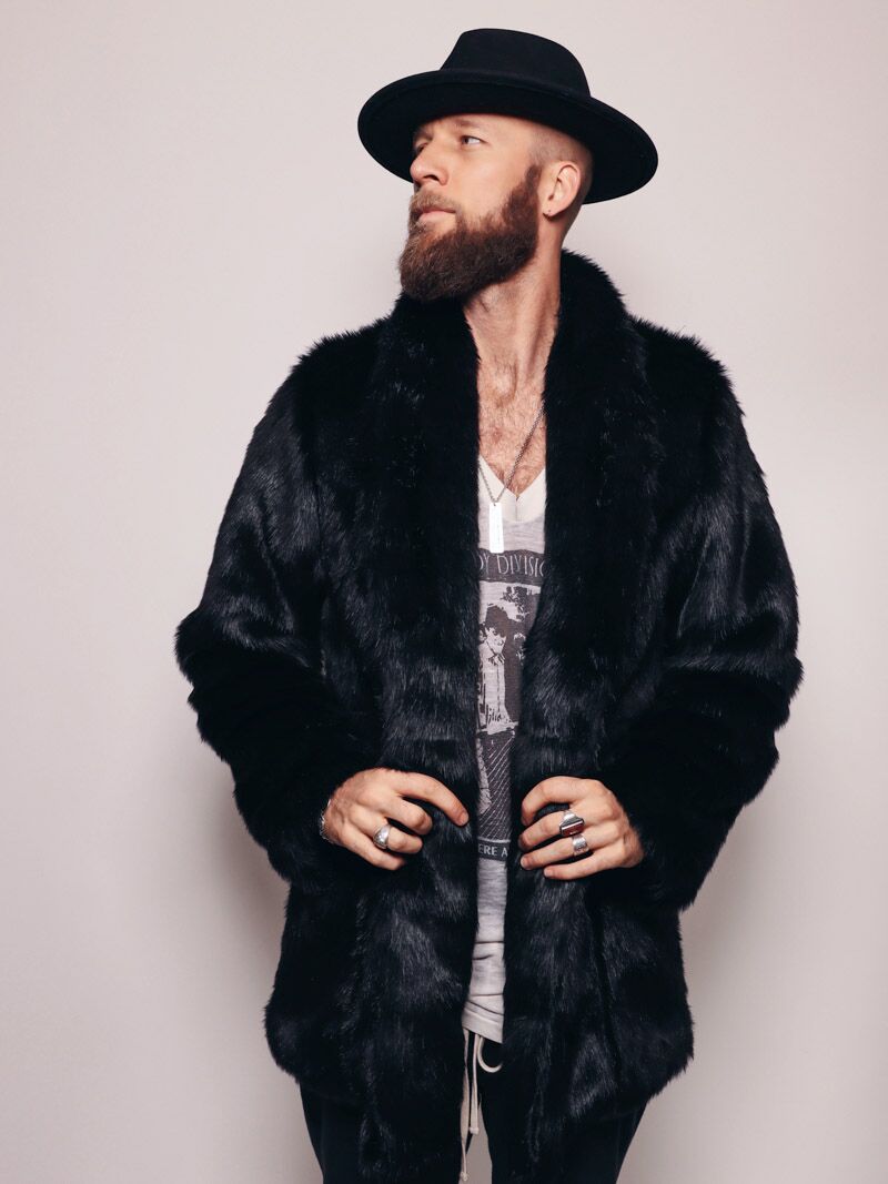 Dude with fedora, beard and mustache looking to one side holding bottom part of the fake fur black panther coat with a collar. 