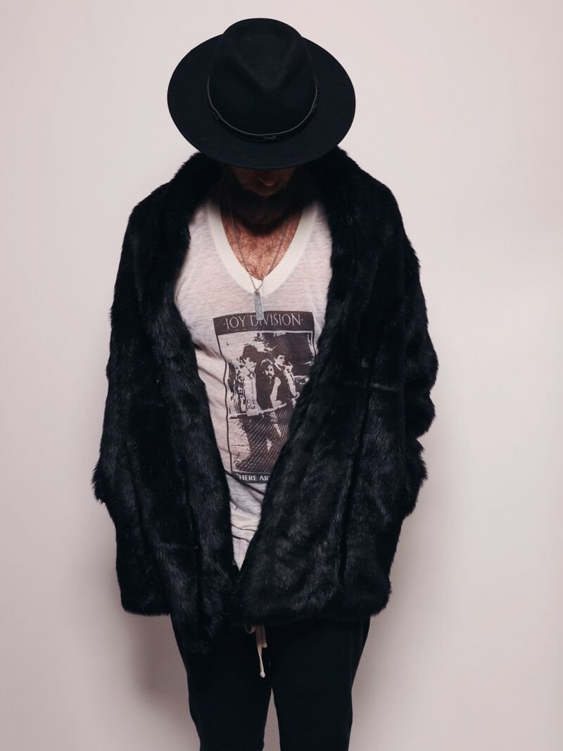 Male model wearing a fedora with his head down and a shadow over his face wearing a panther fake fur coat that is black, with his hands in the pockets. 