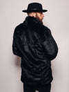 Guy in a fedora with a beard and sideburns with his back to the camera and looking to the left wearing a hoodless black panther fake fur jacket. 