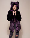 Blonde female model wearing midnight wolf dark purple faux fur coat with hood and ears holding the neck with both hands and staring into camera with slight smile on her face. 