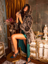 Model holding perfume bottle towards her face with one leg up on a small round table  in a fancy room while wearing a fake fur grey wolf coat with the hood on. 