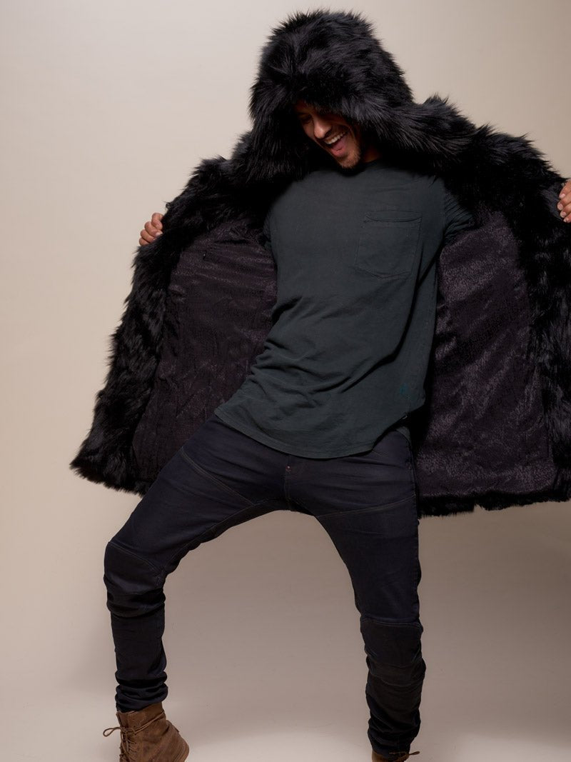 A happy male model is smiling with his mouth open, holding the faux fur black wolf coat open with both hands, the hood on, showcasing the liner while raising one knee as if he’s doing a dance move.