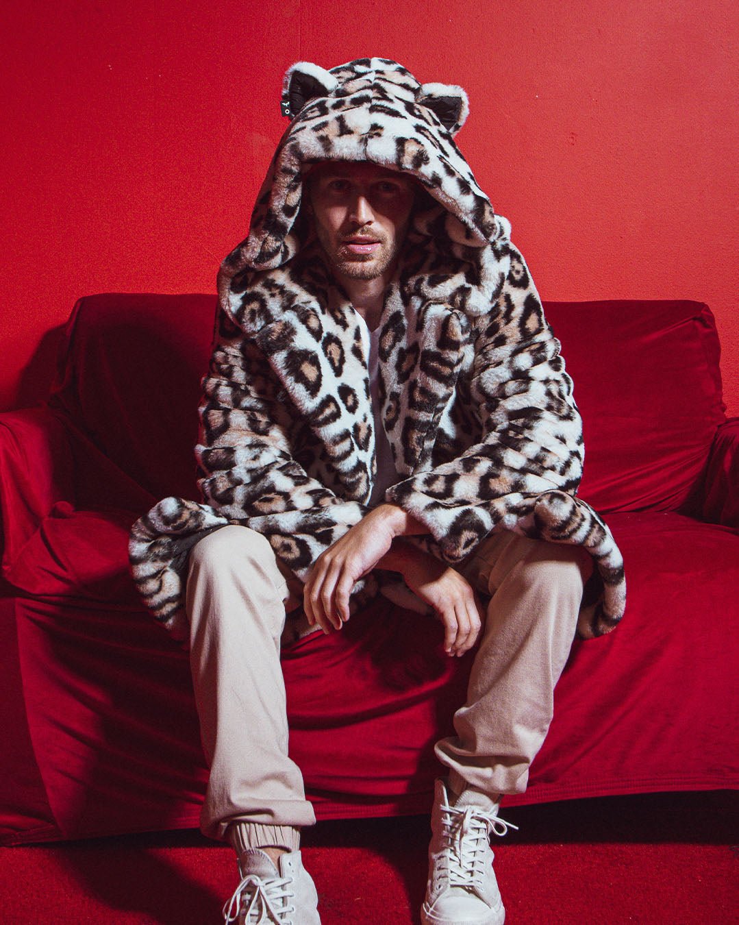 Man in red room, on red couch, leaning forward with arms crossed over knees in Classic Men&#39;s Long Faux Fur Coat in Arabian Leopard Print, hood up.