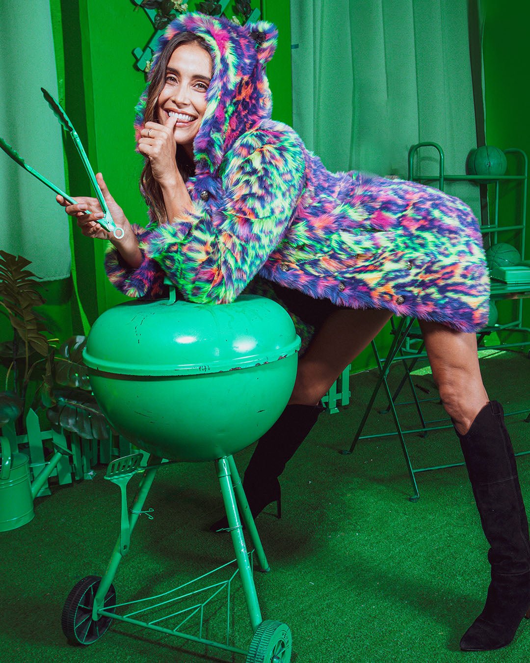 Girl leans over a green BBQ on an all-green patio, wearing the Neon Disco Leopard faux fur jacket.