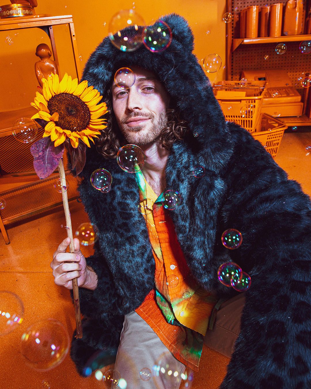 Dude in an orange room full of bubbles, holding a sunflower, wearing the Classic Indigo Leopard Collector Edition Faux Fur Coat with hood and ears up.