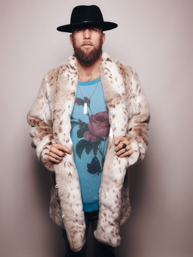 A guy in a black fedora, facing the camera and holding the bottom of his coat, is wearing a jacket made of faux fur called the Siberian snow leopard coat.