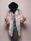 A bearded male model wearing a fedora, with his head down and hands making a 'come here' gesture, is showcasing a faux fur snow leopard collared coat.