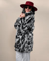 Dark-haired woman in red fedora and sunglasses, wearing Collared Himalayan Snow Leopard Fake Fur Coat closed at neck, smiling gently at camera.