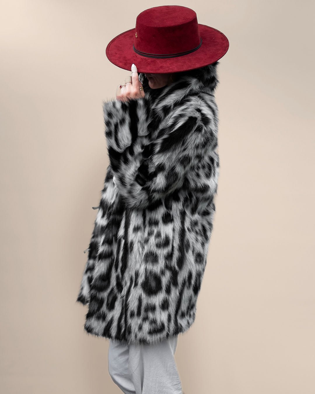 Girl in red fedora covering her face, holding the rim to one side, wearing Collared Himalayan Snow Leopard Faux Fur Jacket. Stylish look.