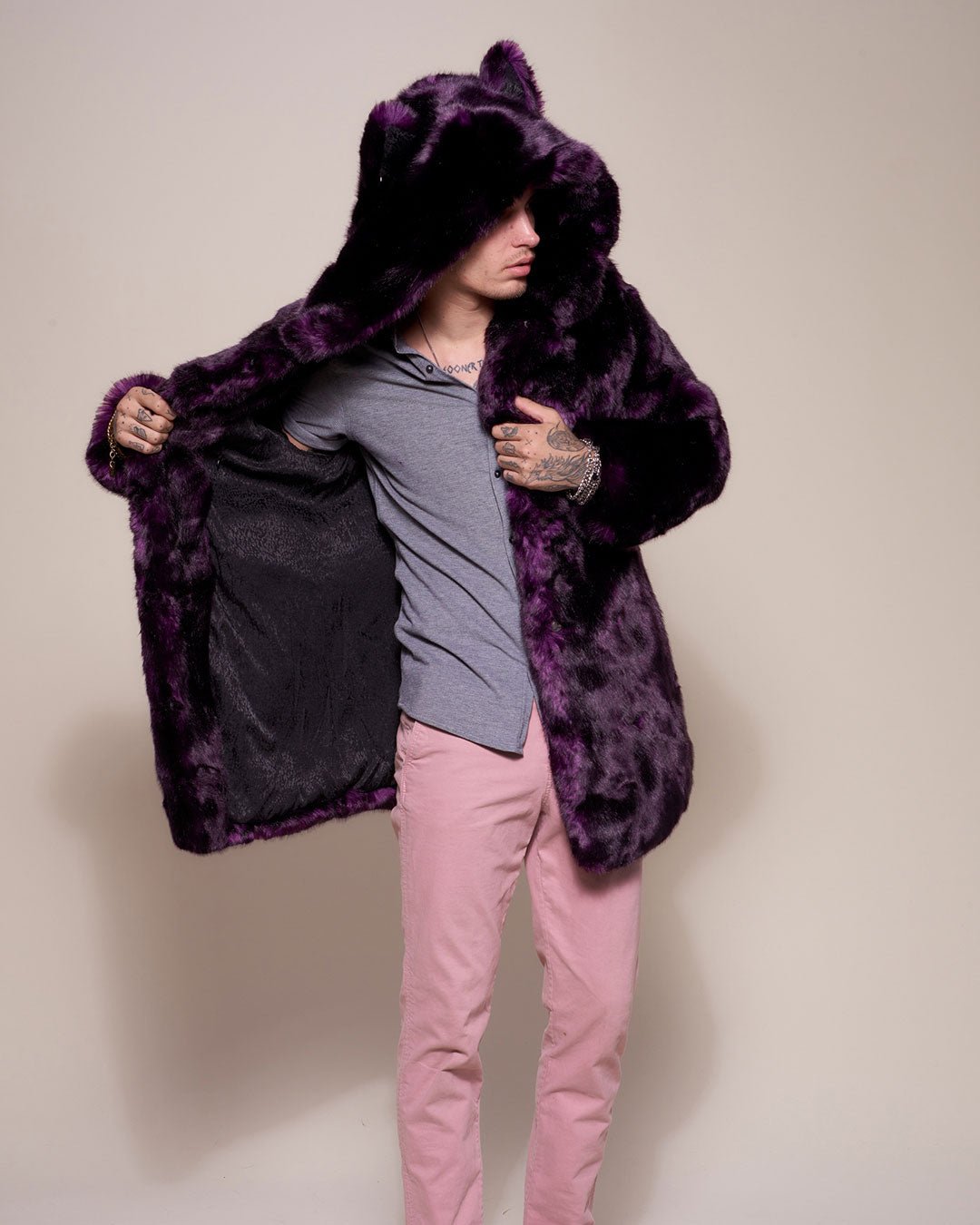 Guy with midnight wolf dark purple artificial fur coat with hood and ears up, turned to one side while holding on side of coat open showing black logo liner. 