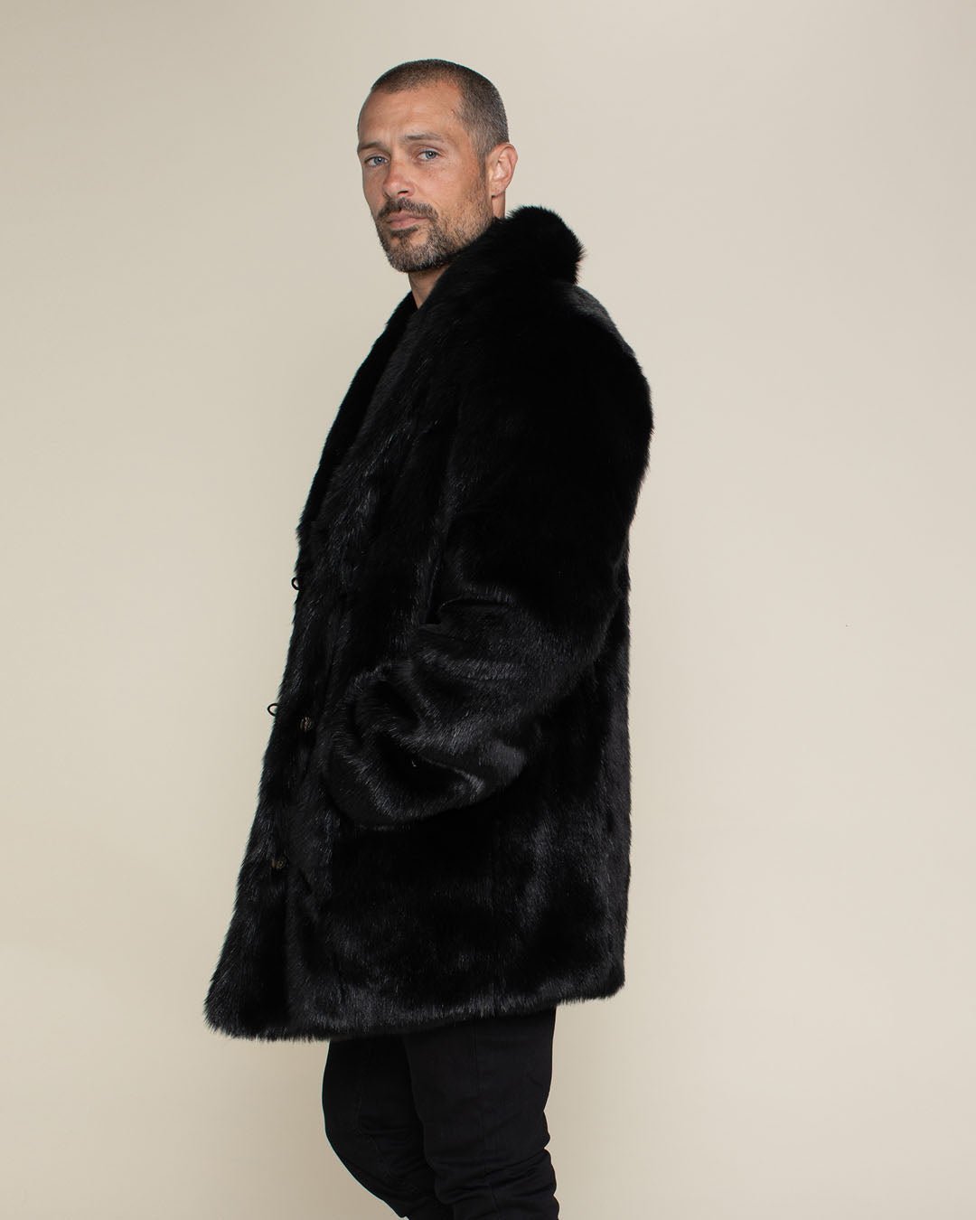 Almost bald dude with short beard and mustache facing the left but looking at the camera wearing collard faux fur black panther jacket with hand in pocket. 