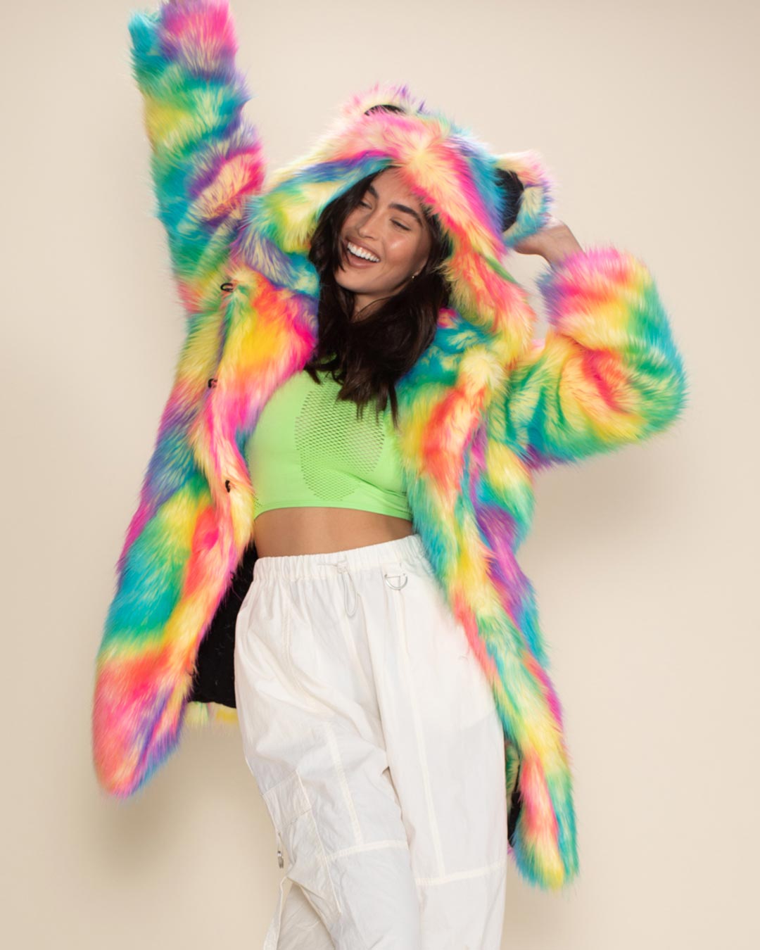 Blissful girl in Rainbow Bear Classic vegan fur jacket, one arm pointing up, other hand on ear, leaning playfully to one side.