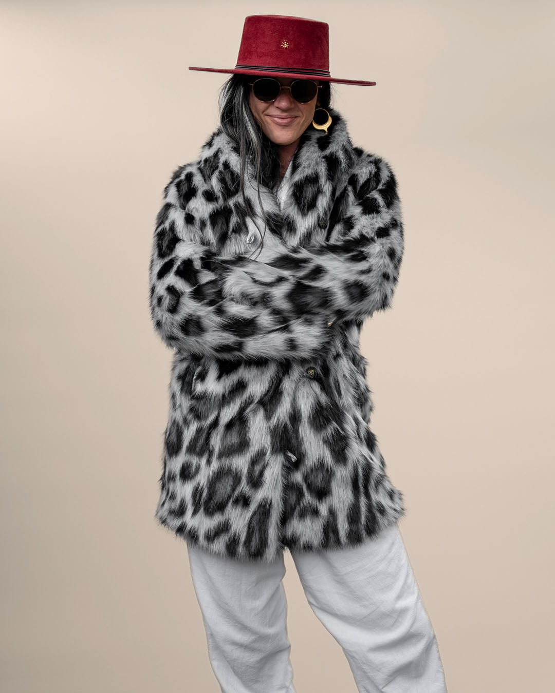 Dark-haired model in red fedora, sunglasses, and Collared Snow Leopard Himalayan Vegan Fur Jacket, gently smiling with arms crossed.