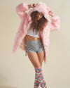 Female model leans with legs crossed, arms up in a stretch, touching the hood of her Flamingo Wolf faux fur coat.