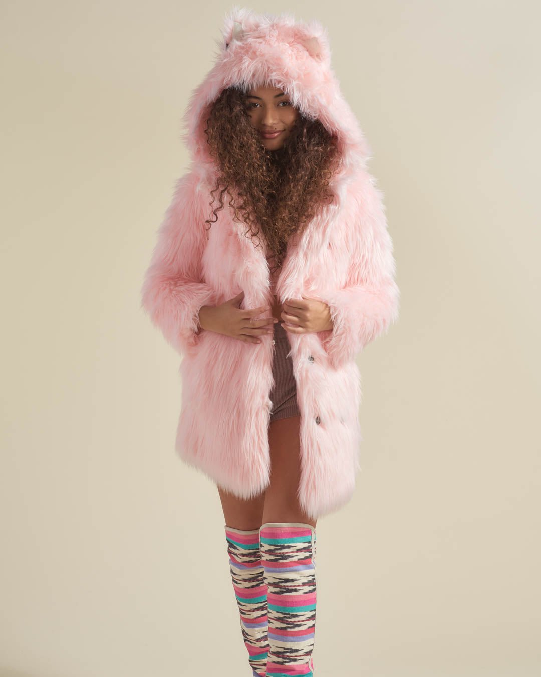 Curly-haired model smiles in a Flamingo Wolf faux fur jacket with hood, ears up, holding lapels, wearing striped knee-highs.