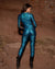 Woman standing facing red rock formations, showing the reflective snakeskin bodysuit from behind