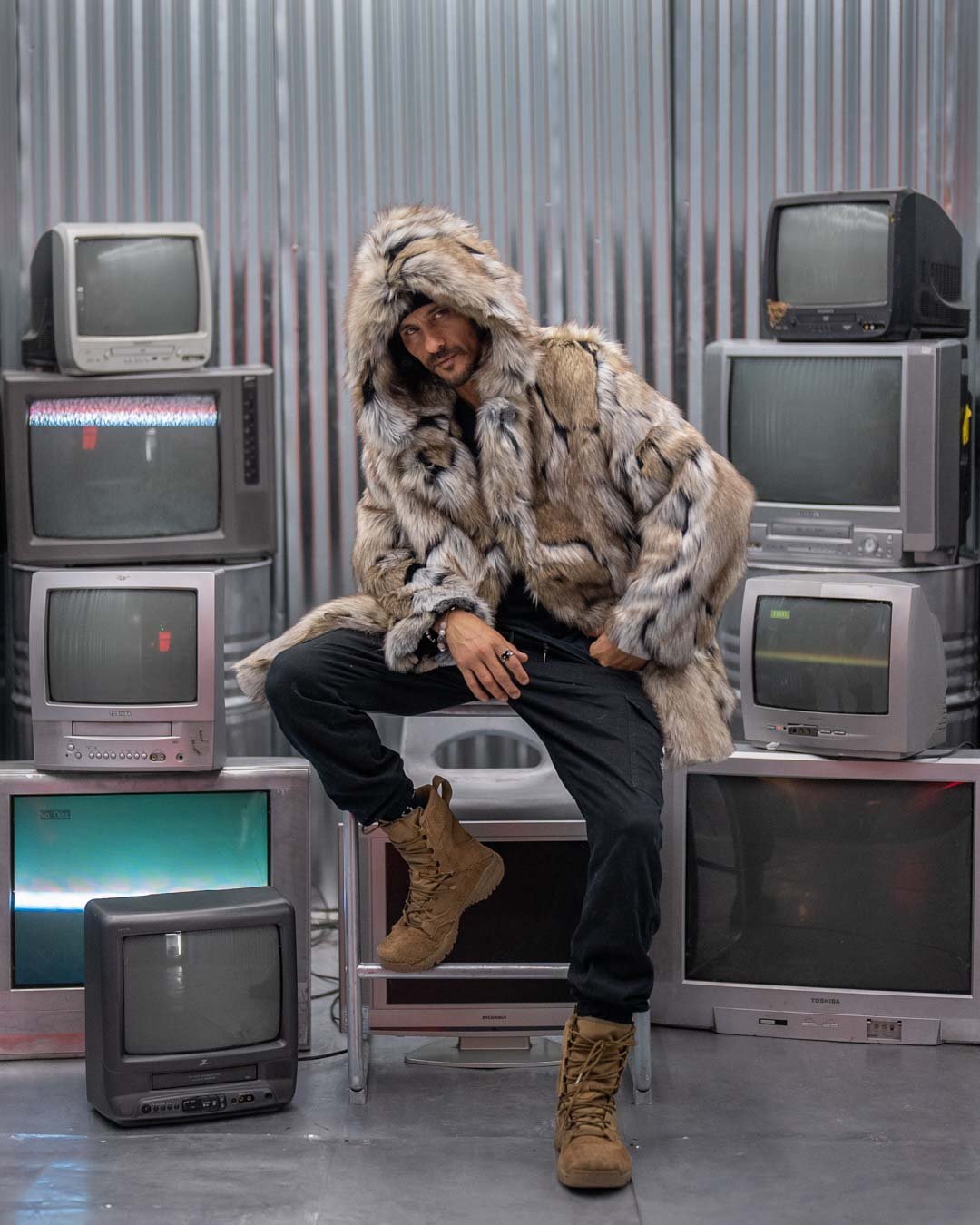 A handsome male model surrounded by old TVs and monitors sits on one, leaning and looking up from under the Hooded Wolverine jacket.