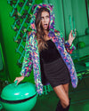 Surprised girl with green tongs holds the lid of a green BBQ on a green patio, wearing her Neon Leopard Cat Disco faux fur jacket.