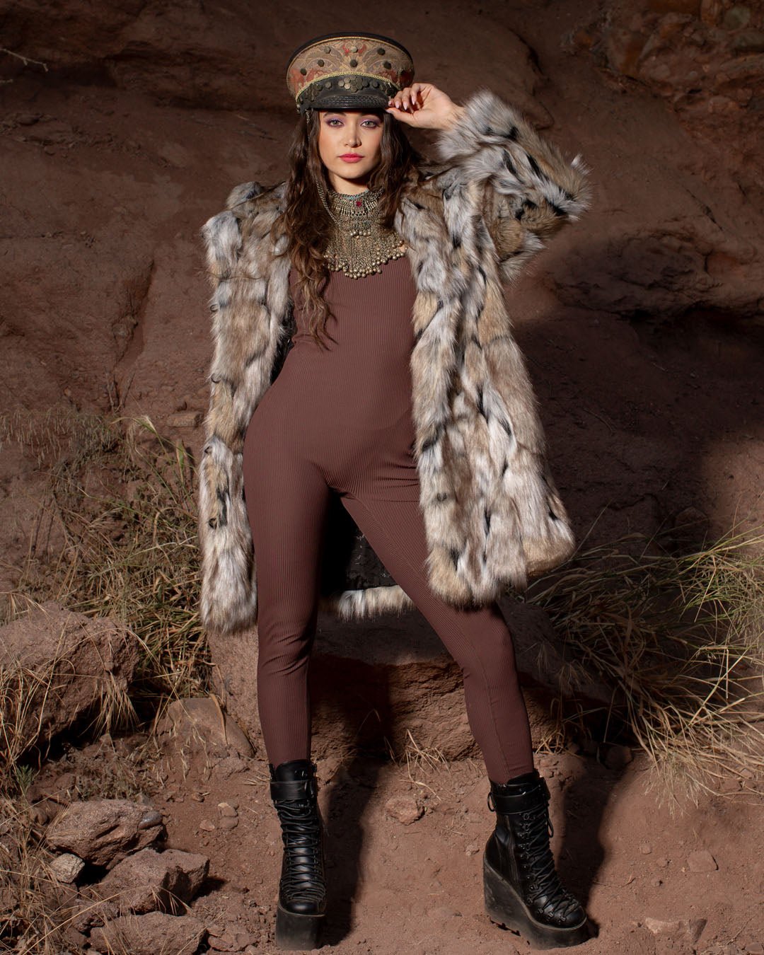 A female model with orange lipstick outdoors, wearing a realistic Hooded Wolverine coat with the hood down, her hand on the bill of a captain's hat, over a brown bodysuit with black combat boots.