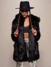 Serious girl model with fedora and braids facing camera wearing wolf coat made of realistic black fur.