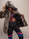 Gal in fake fur hooded grey wolf coat, doing a dance move while holding one side of coat open exposing black logo liner.