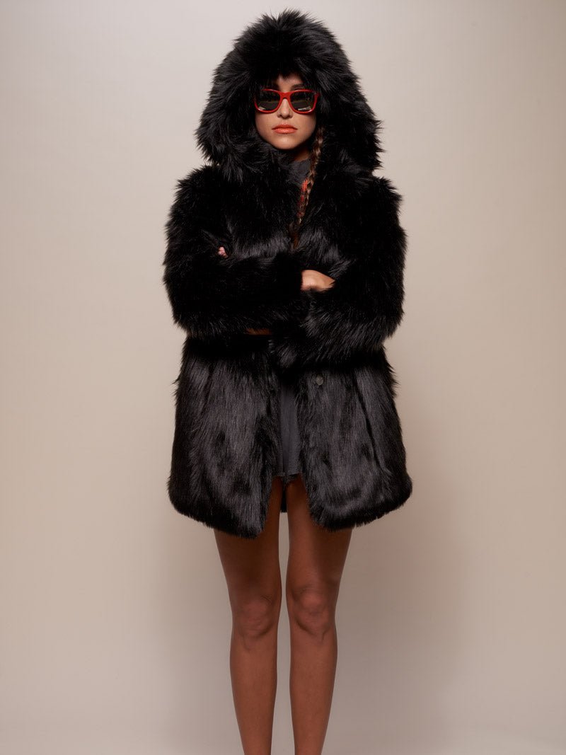 A serious girl wearing red sunglasses and a hooded black wolf faux fur coat with the hood up, her arms crossed over her chest.