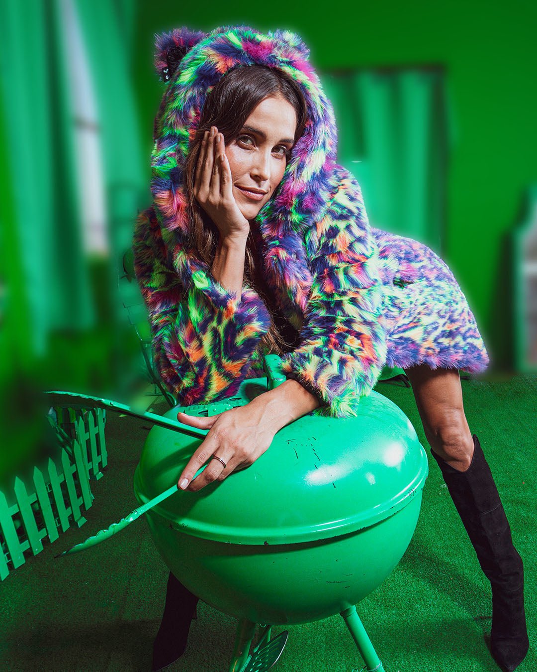 Sweet-faced girl leans on a green BBQ with green tongs on a green patio, wearing the Neon Disco Leopard Cat faux fur coat.