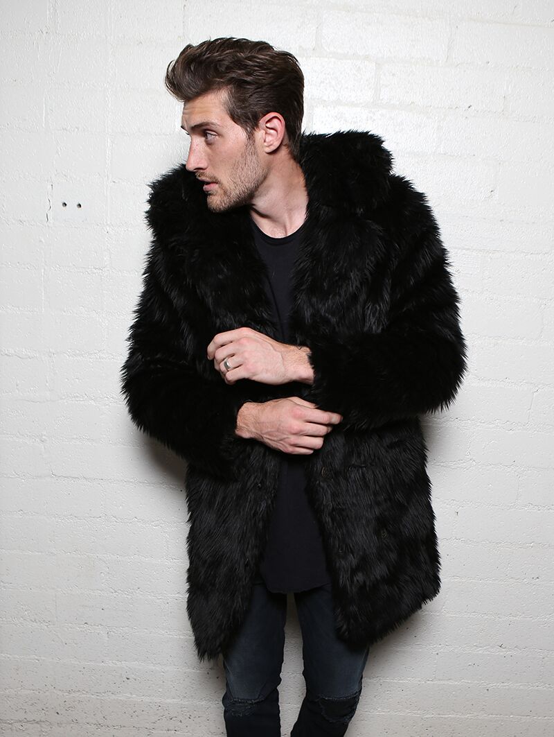 Male model with short brown hair looking to the right wearing vegan fur black wolf coat, with arms almost crossed in front of him. 