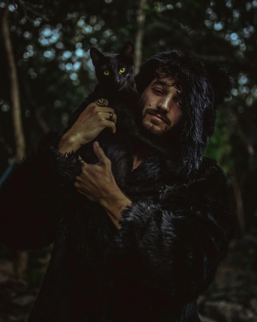 Mustache and bearded male model holding a black kitten in a forest while wearing fake fur black panther jacket